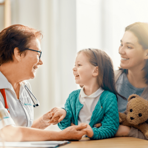 A happy, smiling woman holding a teddy bear with a happy toddler, who is smiling and looking at another smiling woman, who is wearing scrubs and has a stethoscope.