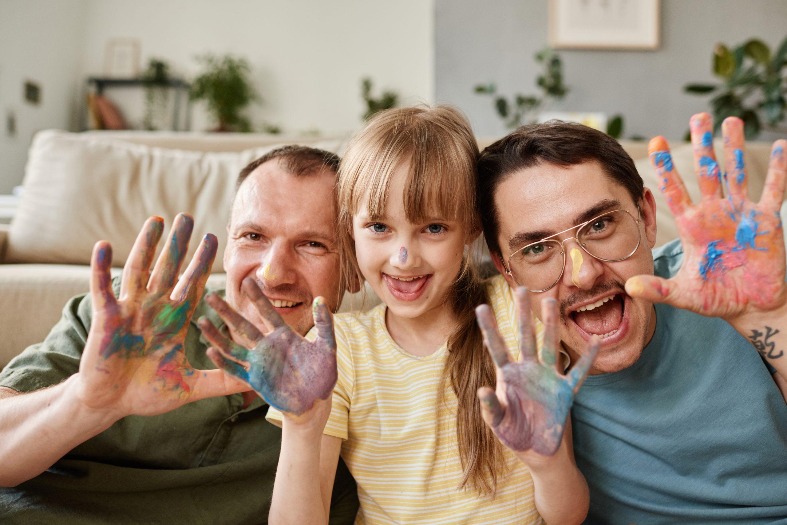 Happy family with child painting at home; Portrait of happy gay family with child showing their painted hands and smiling at camera