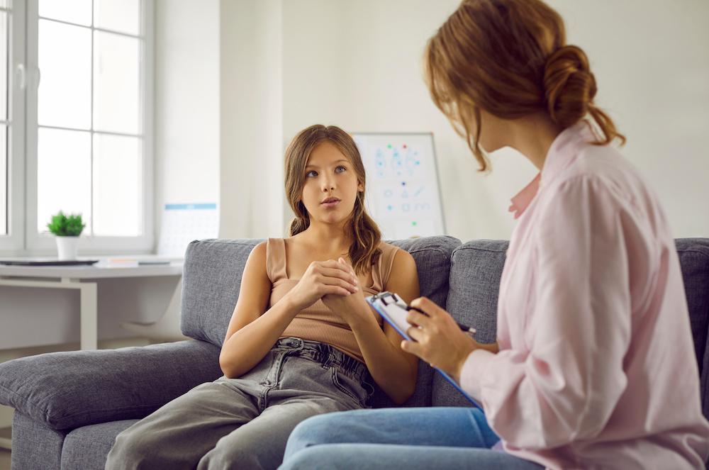 An Outpatient Therapy Session with a psychologist holding a clipboard while listening to the child. Professional therapist working with teenage child. Adolescent girl sitting on sofa and answering questions that school psychologist asks her.